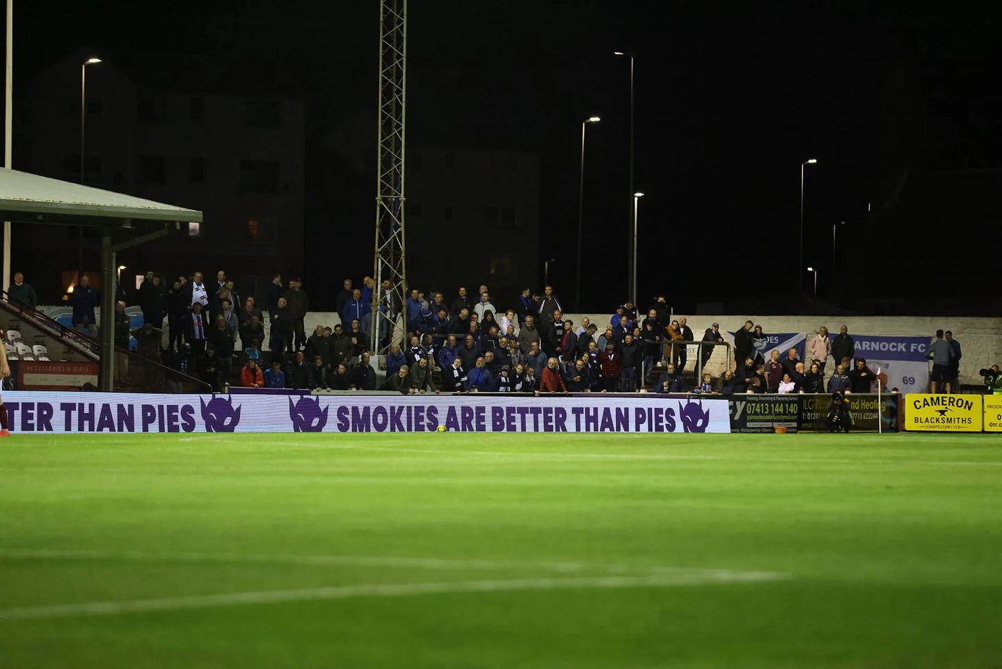 A floodlit football match with a pitchside LED board reading 'Smokies are better than pies' next to the Purple Imp logo
